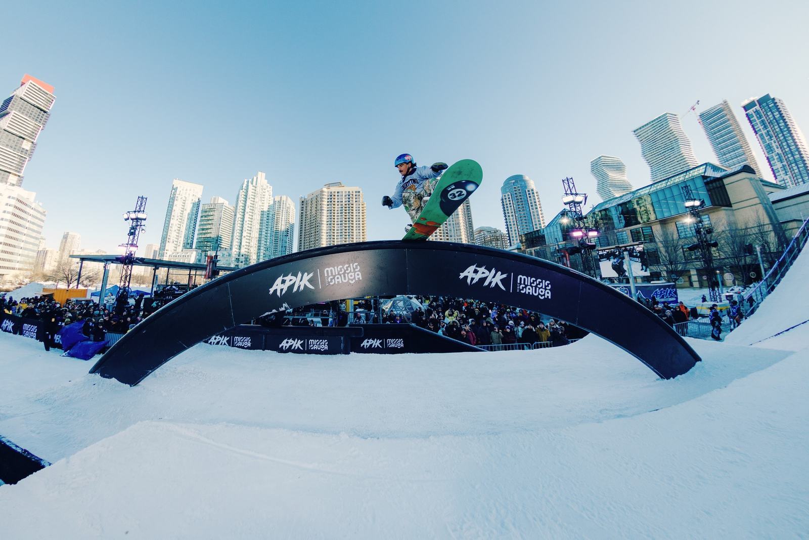 A snowboarder rides the APIK rainbow rail in downtown Mississauga, towers and a packed crowd behind