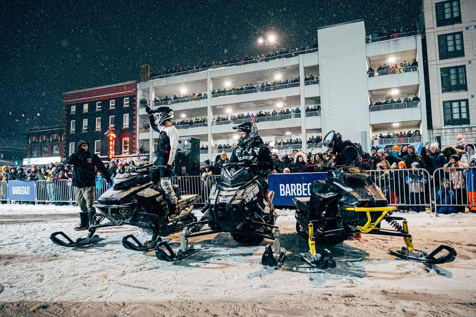 Snowmobile freestyle riders line up in falling snow at Barbegazi Ottawa, crowds packed on four levels of a parking structure