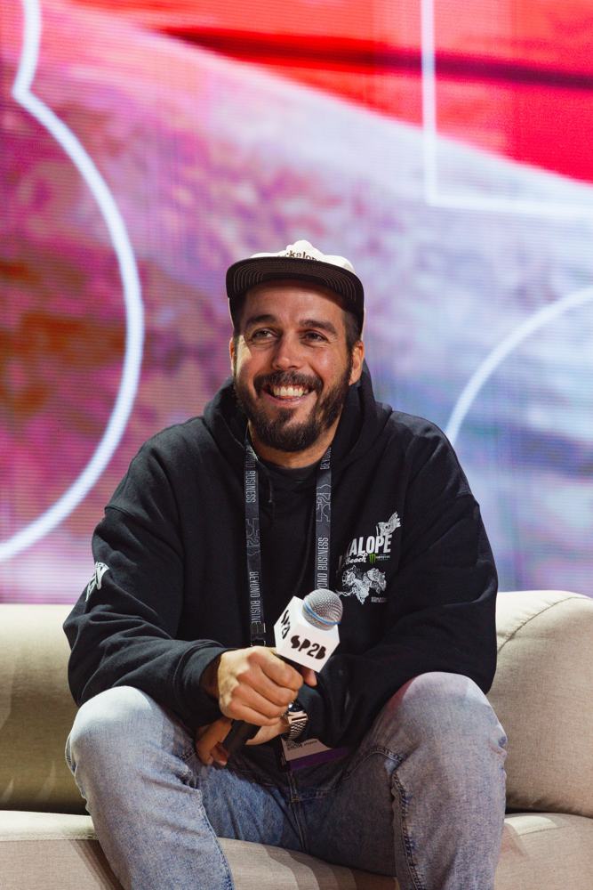 Micah laughing on the SP2B couch, mic in hand, against a red and pink screen
